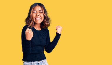 Genç, güzel, melez, günlük kıyafetler giyen, mutlu ve heyecanlı, kolları havada, gülümseyen ve başarı için bağıran, kazanan bir jest yapan bir kadın. kutlama konsepti. 