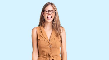 Genç, sarışın, sıradan kıyafetler giyen, gözlüklü, dilini dışarı çıkaran güzel bir kadın. Komik bir ifadeyle mutlu. duygu kavramı. 