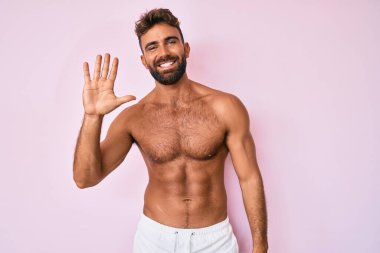 Genç İspanyol adam üstsüz duruyor ve gülümserken beş numarayı işaret ediyor. Kendinden emin ve mutlu.. 