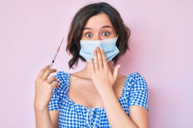 Tıbbi maske takan genç ve güzel bir kadın elinde şırıngayla ağzını eliyle kapatıyor, şok geçiriyor ve hata yapmaktan korkuyor. şaşırmış ifade 