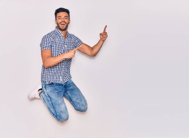 Genç, yakışıklı, sıradan kıyafetler giyen, mutlu gülümseyen bir İspanyol. Yüzünde tebessümle sıçrayıp, izole beyaz arka planda parmaklarını yana doğru gösteriyor.