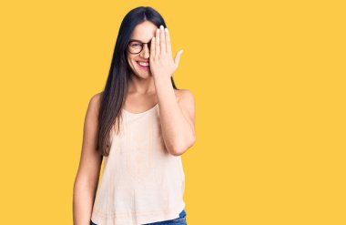 Gündelik kıyafetler ve gözlük takan genç beyaz bir kadın bir gözünü eliyle kapatıyor, yüzünde kendine güvenen bir gülümseme ve şaşırtıcı bir duygu var.. 