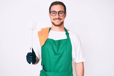 Önlük takan genç yakışıklı adam elinde tuvalet fırçası tutarken pozitif ve mutlu görünüyor ve kendine güvenen bir gülümsemeyle dişlerini gösteriyor. 