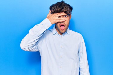 Gündelik kıyafetler giyen genç İspanyol adam şok içinde yüzünü ve gözlerini eliyle kapatıyor, parmaklarından korkuyla bakıyor. 