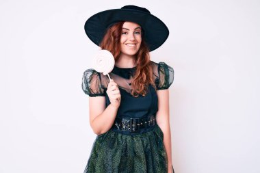 Cadı cadılar bayramı kostümü giymiş genç bir kadın elinde lolipop tutarken pozitif ve mutlu görünüyor. Kendine güvenen bir gülümsemeyle dişlerini gösteriyor. 