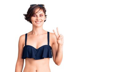 Kısa saçlı, bikini giyen, gülümseyen, kameraya göz kırpan, zafer işareti yapan güzel bir genç kadın. İki numara.. 