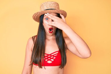 Bikini giyen, şapkalı, güzel beyaz bir kadın. Şoka girmiş, yüzünü ve gözlerini eliyle kapatmış, parmaklarından korkuyla bakıyor. 