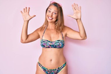 Orta yaşlı İspanyol kadın bikini giyer ve parmaklarını 10 numaraya doğrultur. Bu arada kendine güvenen ve mutlu gülümser.. 