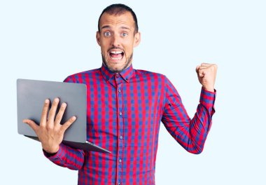 Bilgisayar laptopuyla çalışan yakışıklı genç adam gururla bağırıyor, zaferi kutluyor ve başarıyı kollarını kaldırarak kutluyor. 