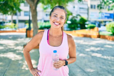 Orta yaşlı sporcu kadın parkta elinde su şişesiyle mutlu bir şekilde gülümsüyor.