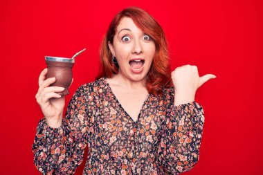 Genç, güzel, kızıl saçlı bir kadın izole edilmiş kırmızı arka planda eş içeceği içerken başparmağını yana uzatıyor ve ağzı açık gülümsüyor.