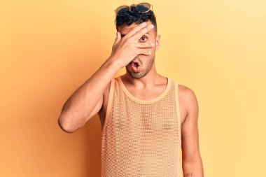 Gündelik kıyafetler giyen genç İspanyol adam şok içinde yüzünü ve gözlerini eliyle kapatıyor, parmaklarından korkuyla bakıyor. 