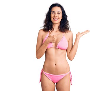 Genç, İspanyol asıllı, bikinili güzel bir kadın avuçlarını gösteriyor ve baş parmaklarıyla iyi bir jest yapıyor, mutlu ve neşeli gülümsüyor. 