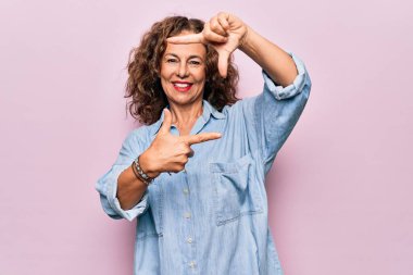 Orta yaşlı güzel bir kadın pembe arka plan üzerinde sıradan bir kot gömlek giyiyor gülümsüyor ve elleri ve parmakları mutlu yüzlü bir çerçeve yapıyor. Yaratıcılık ve fotoğrafçılık kavramı.