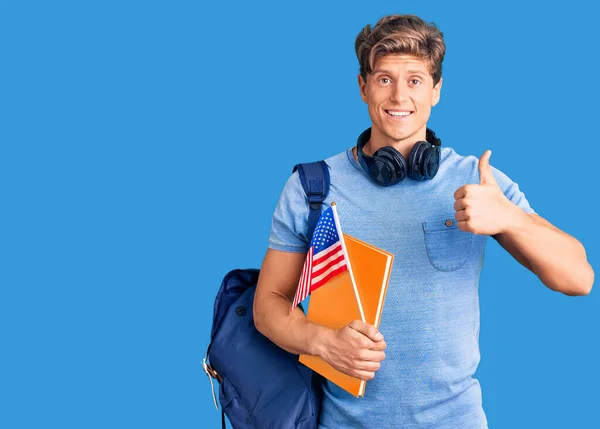 年轻英俊的男子 背着学生背包和耳机 拿着书本和美国国旗 开心而积极地微笑着 竖起大拇指做得出色 签了字 — 图库照片
