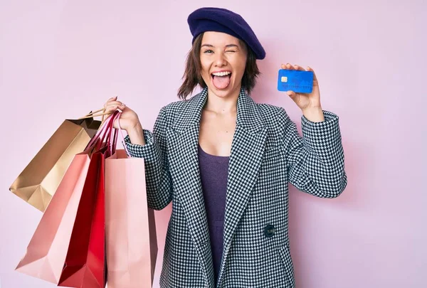 年轻美丽的女人头戴贝雷帽 提着购物袋和信用卡 带着滑稽的表情伸出舌头 高兴极了 — 图库照片