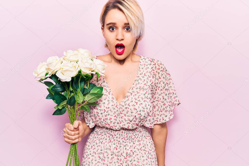 Young beautiful blonde woman holding flowers scared and amazed with open mouth for surprise, disbelief face 