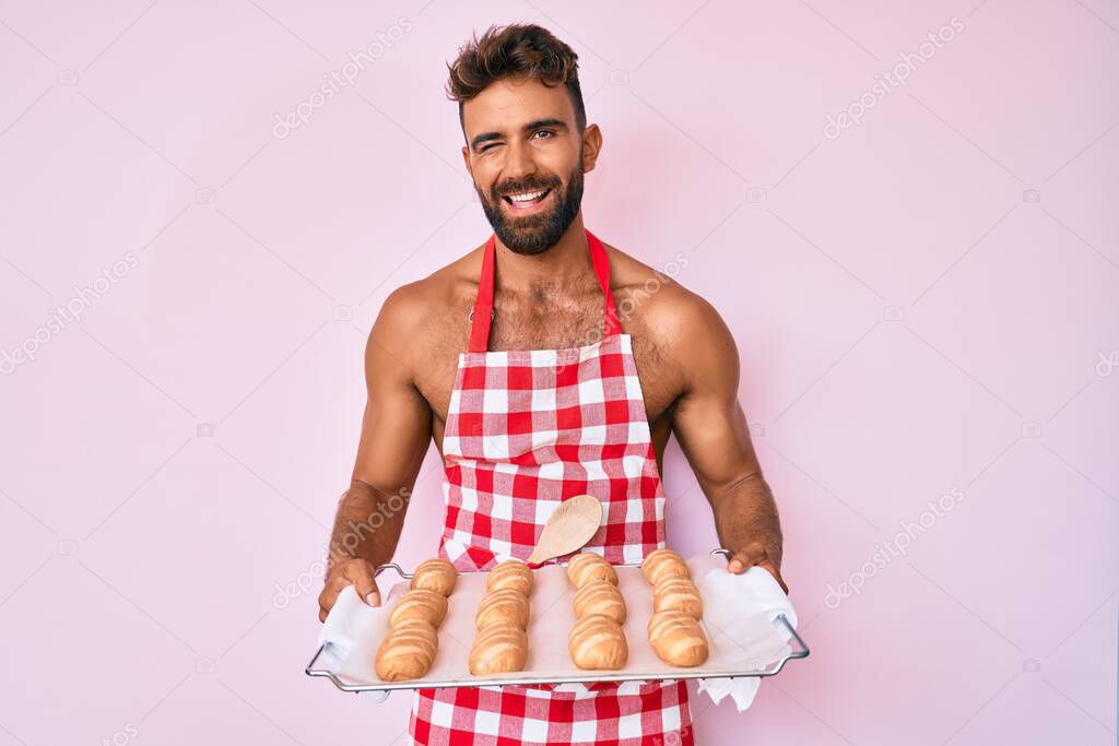 Young hispanic man shirtless wearing baker uniform holding homemade bread winking looking at the camera with sexy expression, cheerful and happy face. 
