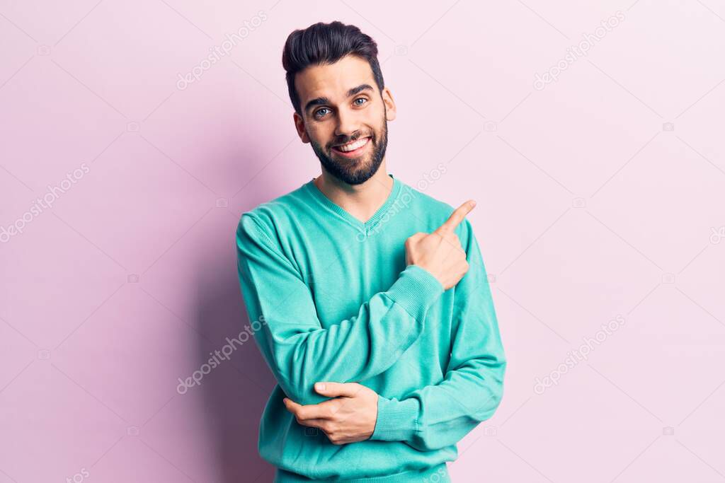 Young handsome man with beard wearing casual sweater smiling cheerful pointing with hand and finger up to the side 