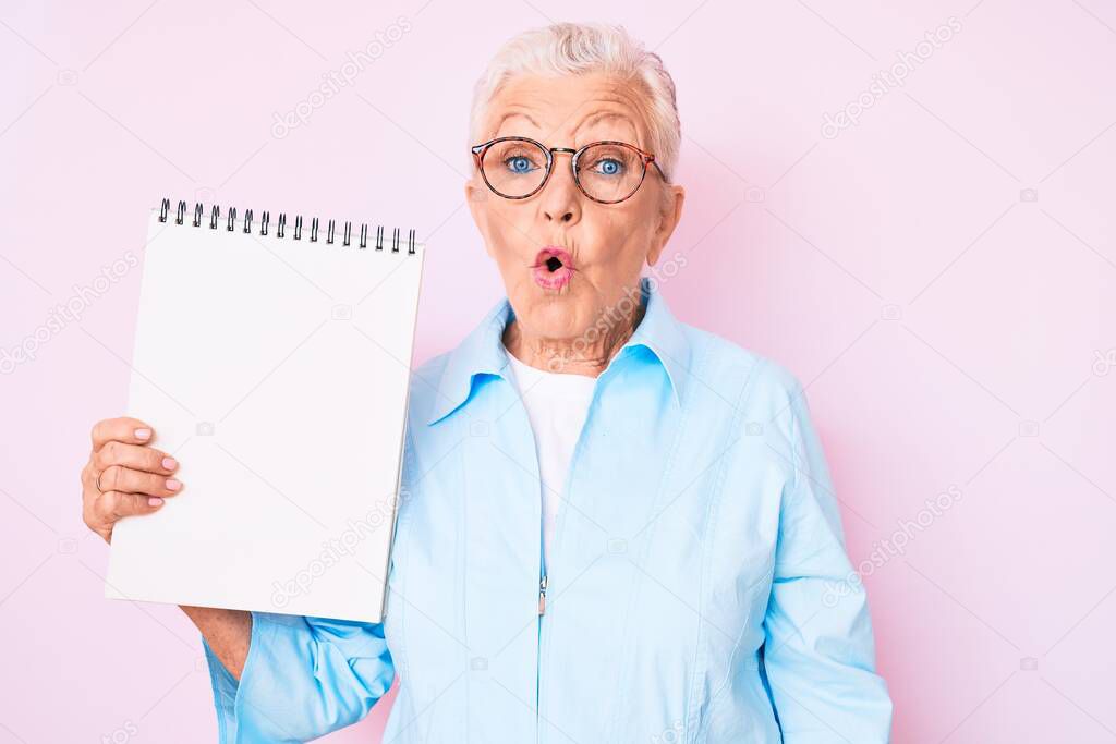 Senior beautiful woman with blue eyes and grey hair holding notebook scared and amazed with open mouth for surprise, disbelief face 