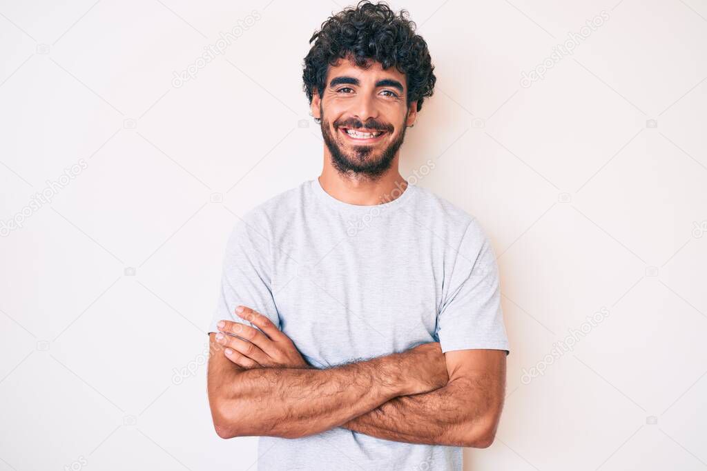 Handsome young man with curly hair and bear wearing casual tshirt happy face smiling with crossed arms looking at the camera. positive person. 