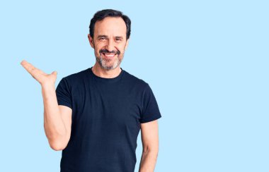 Orta yaşlı yakışıklı bir adam sıradan tişörtler giyiyor neşeli bir şekilde gülümsüyor ve avuç içiyle kameraya bakıyor.. 