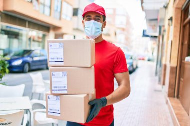 Üniforma giyen ve Coronavirus koruması takan genç bir teslimatçı. Sağlık maskesi. Kasaba sokaklarında karton paketler tutuyor..