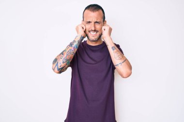 Dövmeli genç yakışıklı adam, gündelik kıyafetler giyiyor kulaklarını parmaklarıyla kapatıyor ve yüksek sesli müzik sesi için rahatsız edici bir ifade takınıyor. sağır kavramı. 