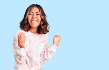 Kravat boyası eşofman üstü giyen genç, güzel, melez bir kadın. Mutlu ve heyecanlı bir şekilde kolları havada, gülümseyerek ve başarı için çığlık atarak kazanan bir jest yapıyor. kutlama konsepti. 