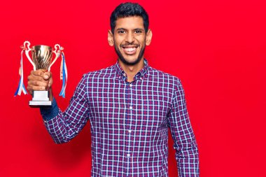 Genç Latin adam zafer kupasını tutuyor. Pozitif ve mutlu görünüyor. Duruyor ve gülümsüyor. 