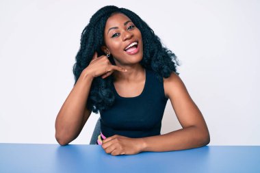 Güzel Afrikalı kadın gündelik giysiler içinde masaya oturmuş gülümsüyor telefonla el ve parmak hareketleri yapıyor telefonda konuşuyor gibi. iletişim kavramları. 