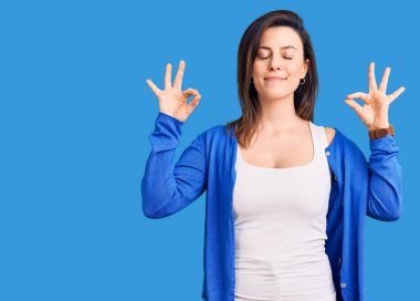 Genç ve güzel bir kadın günlük kıyafetler giyip rahatlıyor ve gözleri kapalı gülümsüyor parmaklarıyla meditasyon hareketi yapıyor. yoga konsepti. 