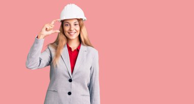 Mimar kasketi takan, gülümseyen, kendine güvenen, el hareketleriyle hareket eden, parmaklarıyla işaret eden ve kameraya bakan güzel bir genç kadın. ölçüm kavramı. 