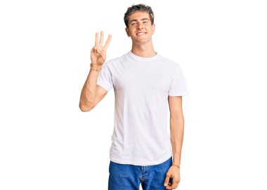Gündelik beyaz tişört giyen genç yakışıklı üç numaralı parmaklarıyla gülümseyerek kendinden emin ve mutlu bir şekilde onu gösteriyor.. 