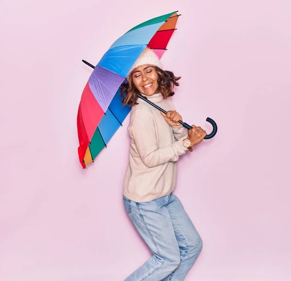 年轻美丽的拉丁女人抱着伞开心地笑着 面带微笑地跳过孤立的粉色背景 — 图库照片