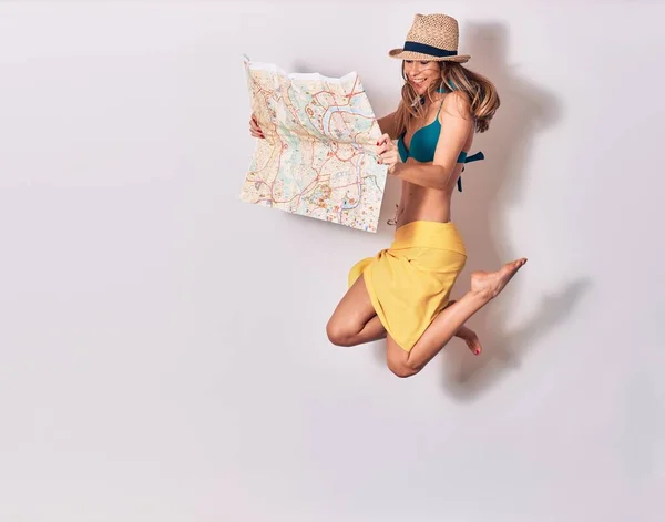 Young beautiful girl wearing bikini and summer hat smiling happy. Jumping with smile on face holding city map over isolated white background