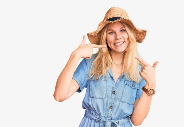 若い美しいブロンドの女性は夏の帽子をかぶって 陽気で笑顔を見せ 指の歯と口で指しています 歯の健康の概念 — ストック写真