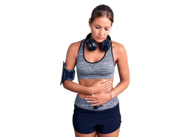 Genç ve güzel İspanyol bir kadın spor kıyafetleri giyiyor ve kulaklık takıyor midesi üstünde çünkü hazımsızlık, acı verici bir hastalık kendini iyi hissetmiyor. Ağrı kavramı. 