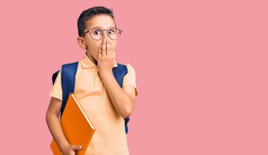 Küçük şirin çocuk okul çantası takıyor ve elinde kitapla ağzını eliyle kapatıyor, şokta ve hata yapmaktan korkuyor. şaşırmış ifade 