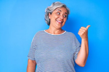 Son sınıf Latin kökenli, gri saçlı, günlük kıyafetler giyen, gülümseyen, gülümseyen ve baş parmağıyla yüzünü gösteren bir kadın.. 