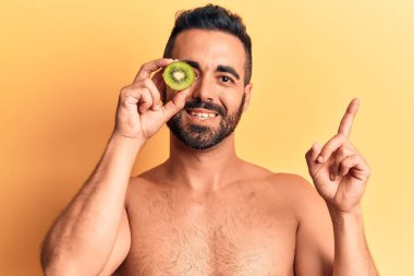 Genç İspanyol adam üstsüz duruyor, elinde kivi var. Gülümsüyor, eli ve parmağı kenarda mutlu bir şekilde işaret ediyor. 