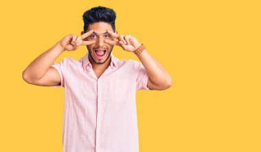 Yakışıklı Latin Amerikalı rahat bir yaz tişörtü giymiş. Barış sembolü yapıyor. Yüzünde parmakları var. 