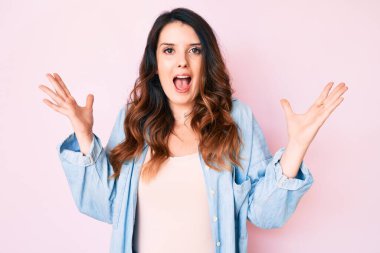 Genç, güzel esmer kadın pembe arka plan üzerine gündelik giysiler giyip zaferi mutlu bir gülümsemeyle ve kazanan yüz ifadesini kaldırarak kutluyor. 