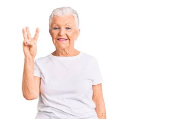 Mavi gözlü, gri saçlı, sıradan beyaz tişörtlü, üç numaralı parmaklarını göstererek gülümseyen, kendinden emin ve mutlu son sınıf güzeli kadın.. 