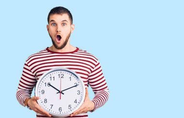 Genç yakışıklı adam büyük saati tutuyor. Korkmuş ve ağzı açık kalmış, şaşırmış ve şaşırmış. 