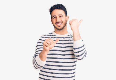 Gündelik kıyafetler giyen genç İspanyol adam arka tarafı işaret ediyor. Elleri havada, kendinden emin bir şekilde gülümsüyor. 