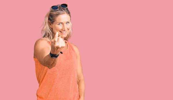 Middle age fit blonde woman wearing casual summer clothes and sunglasses beckoning come here gesture with hand inviting welcoming happy and smiling