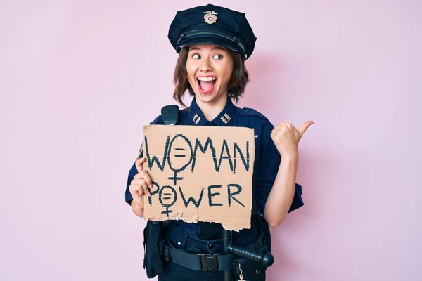 Young beautiful girl wearing police uniform holding woman power banner pointing thumb up to the side smiling happy with open mouth