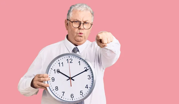 Senior grey-haired man wearing business clothes and holding clock pointing with finger to the camera and to you, confident gesture looking serious