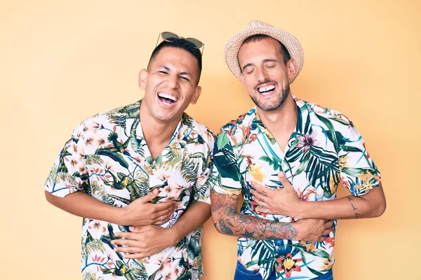 Young gay couple of two men wearing summer hat and hawaiian shirt smiling and laughing hard out loud because funny crazy joke with hands on body.